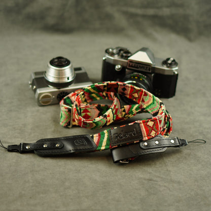 A camera strap with a colorful geometric pattern and a black leather section, displayed in front of two vintage cameras on a textured gray surface.