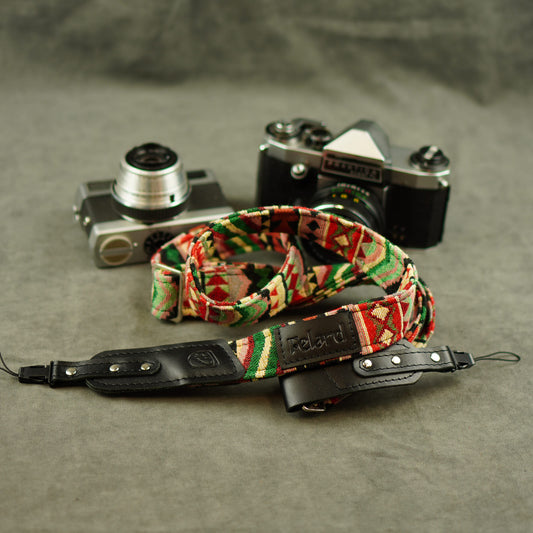 A camera strap with a colorful geometric pattern and a black leather section, displayed in front of two vintage cameras on a textured gray surface.