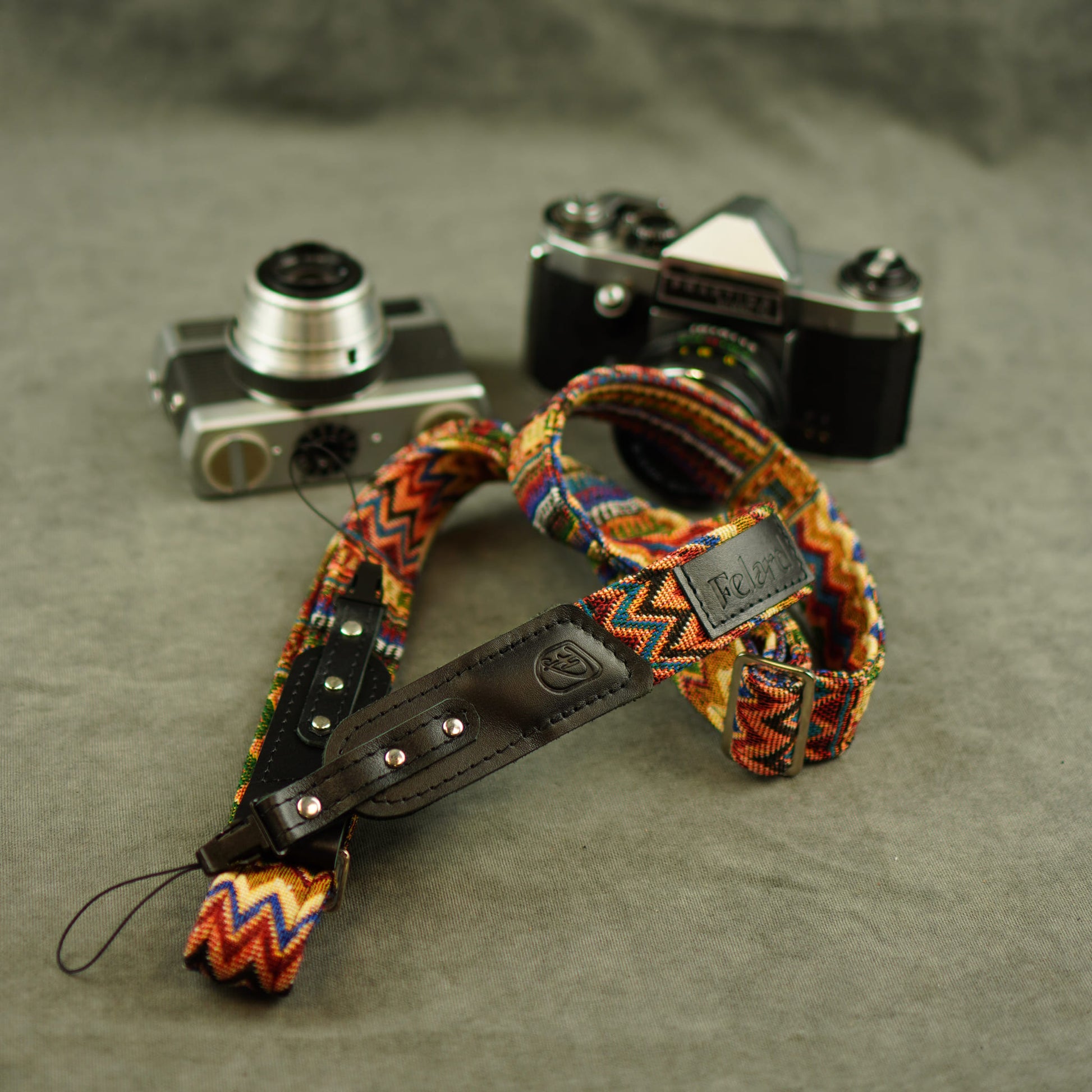 A colorful woven camera strap with a black leather section and silver rivets, displayed on a gray textured surface next to two vintage cameras.