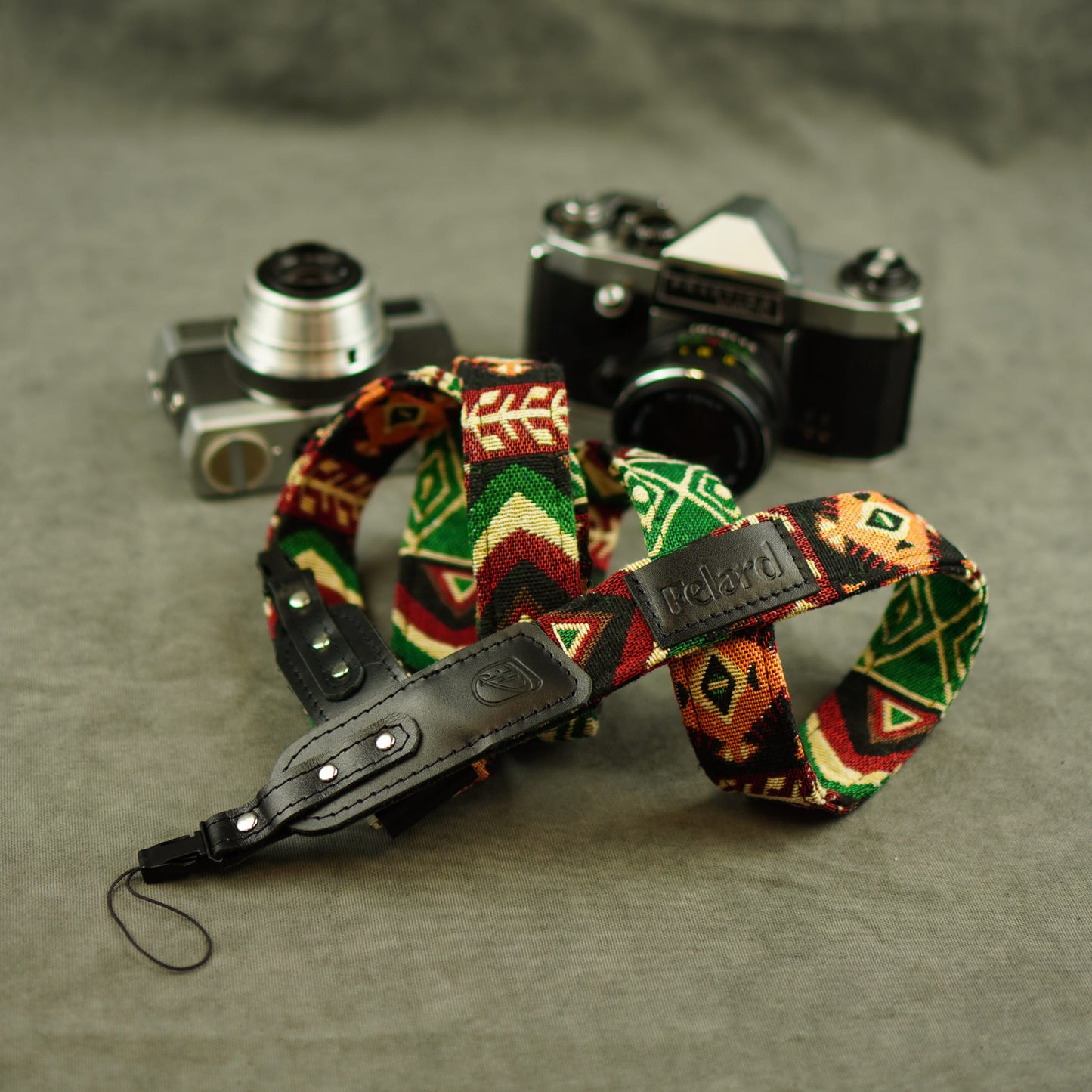 Patterned camera strap with a black leather wristlet and a visible 'Felard' tab, arranged in front of two vintage cameras on a textured gray surface.