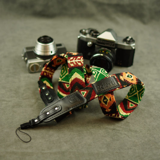 Patterned camera strap with a black leather wristlet and a visible 'Felard' tab, arranged in front of two vintage cameras on a textured gray surface.