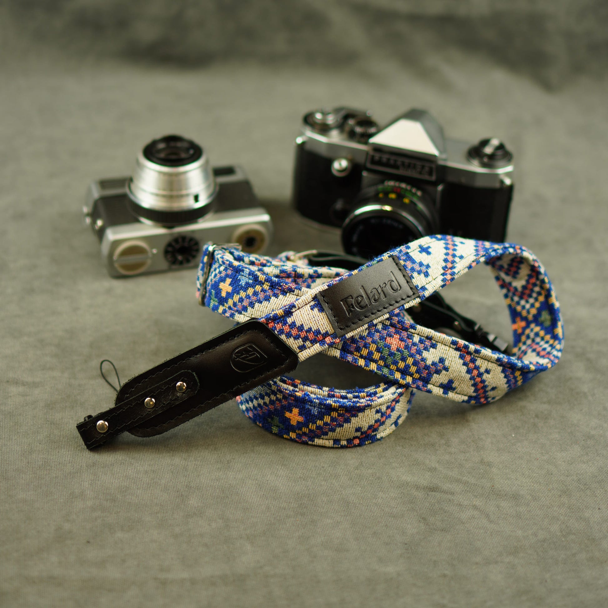 Camera strap with a blue, white, and orange geometric pattern and a black leather displayed in front of two vintage cameras on a textured gray surface.