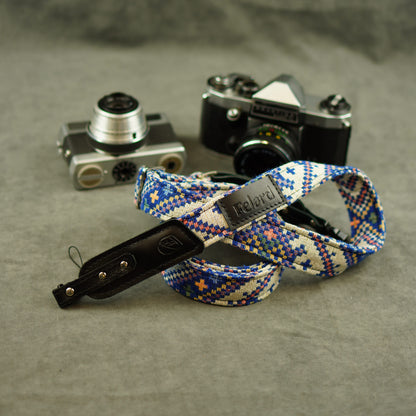 Camera strap with a blue, white, and orange geometric pattern and a black leather displayed in front of two vintage cameras on a textured gray surface.