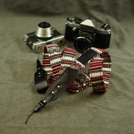 A patterned camera strap with red, black, and white geometric designs and a black leather section, displayed in front of two vintage cameras on a textured gray fabric background.