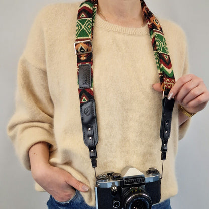 A person holds a vintage-style camera with a patterned fabric strap featuring green, red, black, and beige geometric motifs and black leather sections.
