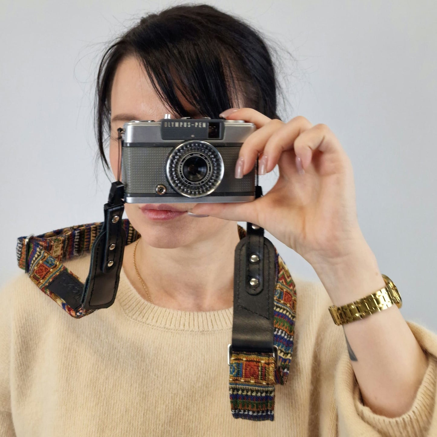 A person holding a vintage silver camera with a colorful patterned leather and fabric strap against a plain light gray background.