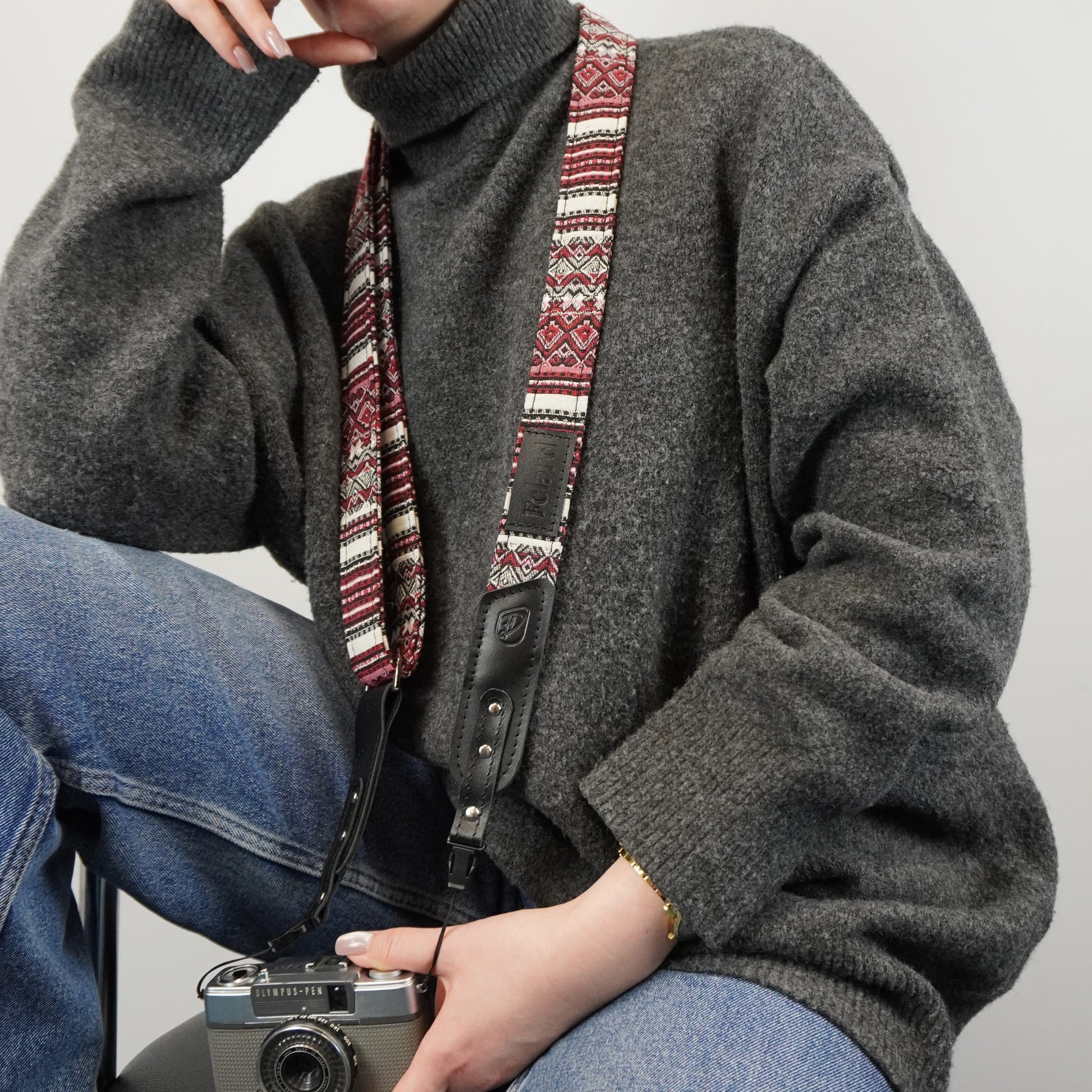 A person holding a vintage camera. The sweater features a prominent woven camera strap with a red, cream, and black geometric pattern and a black leather buckle.