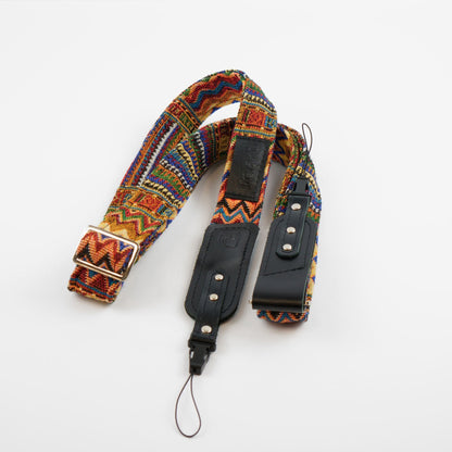 A camera strap featuring a vibrant, multicolored geometric woven pattern with black leather accents and silver-tone hardware, displayed on a clean white background.
