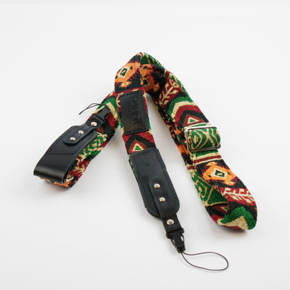A woven camera strap with a vibrant Southwestern geometric pattern in green, red, orange, and black, featuring black leather ends