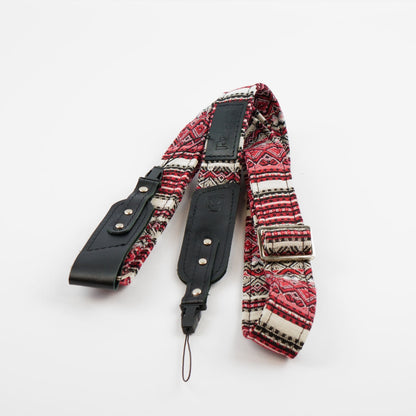 A camera strap featuring a red, black, and white geometric patterned fabric body with black leather accents and silver-tone metal rivets, displayed on a clean white background.