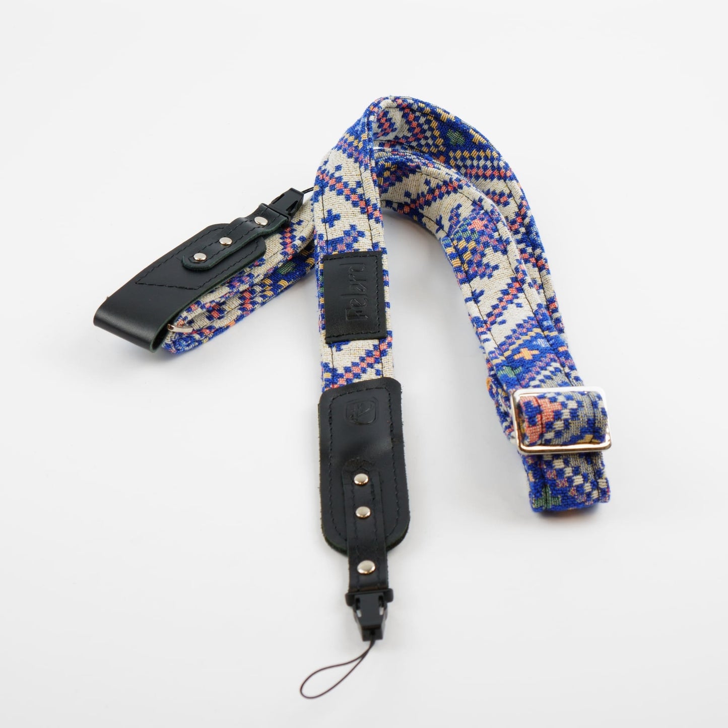 A woven camera strap with a geometric pattern in blue, red, and yellow on a cream background, featuring black leather accents and a silver metal buckle.