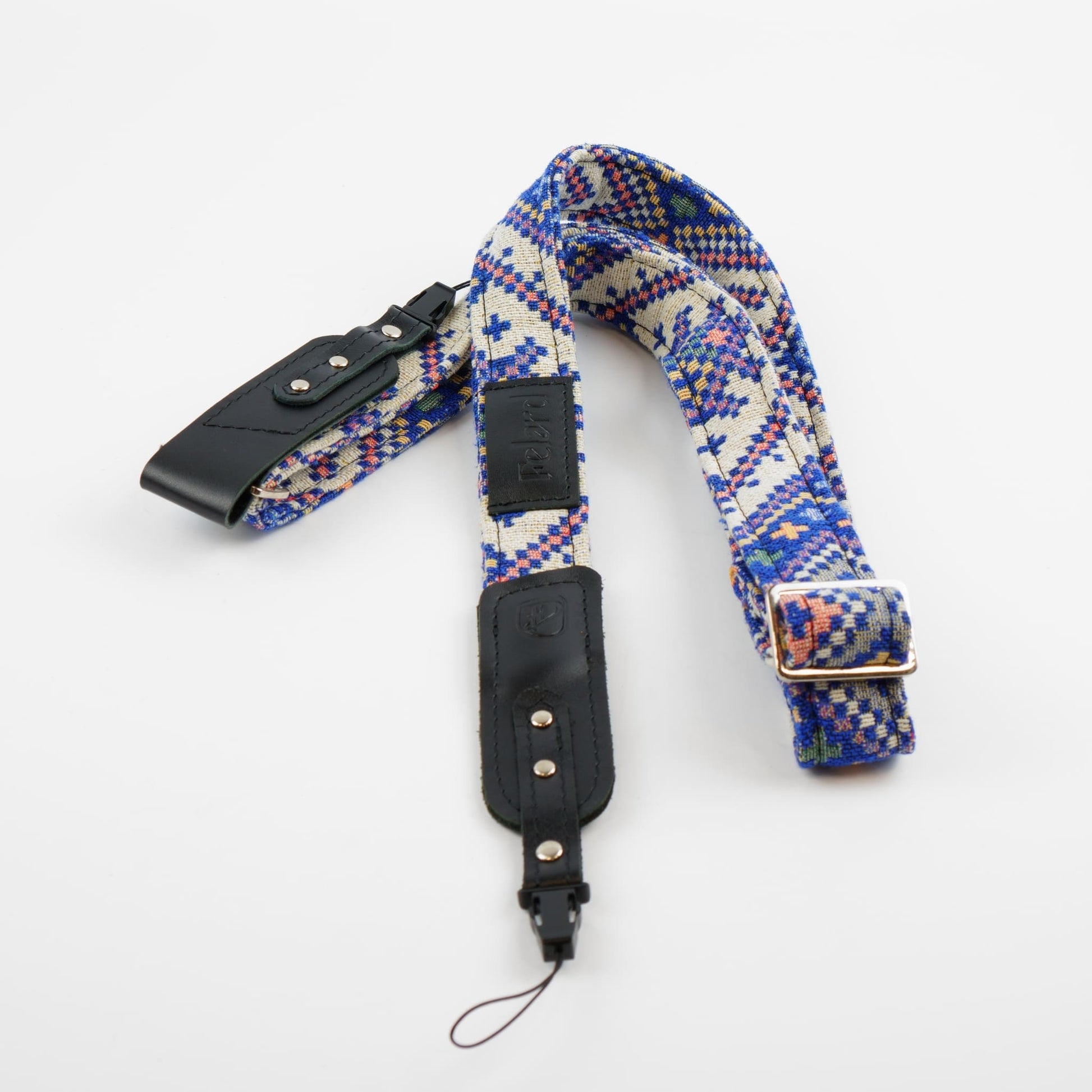 A woven camera strap with a geometric pattern in blue, red, and yellow on a cream background, featuring black leather accents and a silver metal buckle.