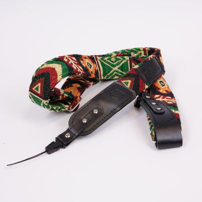 A camera strap featuring a colorful geometric pattern with green, red, orange, and beige, accented by black leather sections and silver metal rivets, displayed on a clean white background.