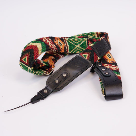 A camera strap featuring a colorful geometric pattern with green, red, orange, and beige, accented by black leather sections and silver metal rivets, displayed on a clean white background.