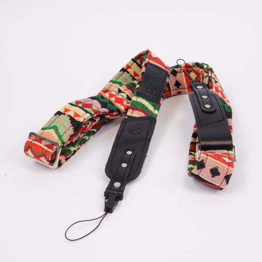 A vintage-style camera strap featuring a colorful geometric pattern in red, green, beige, and black, with black leather webbing and silver-tone metal buckles, laid flat on a white background.
