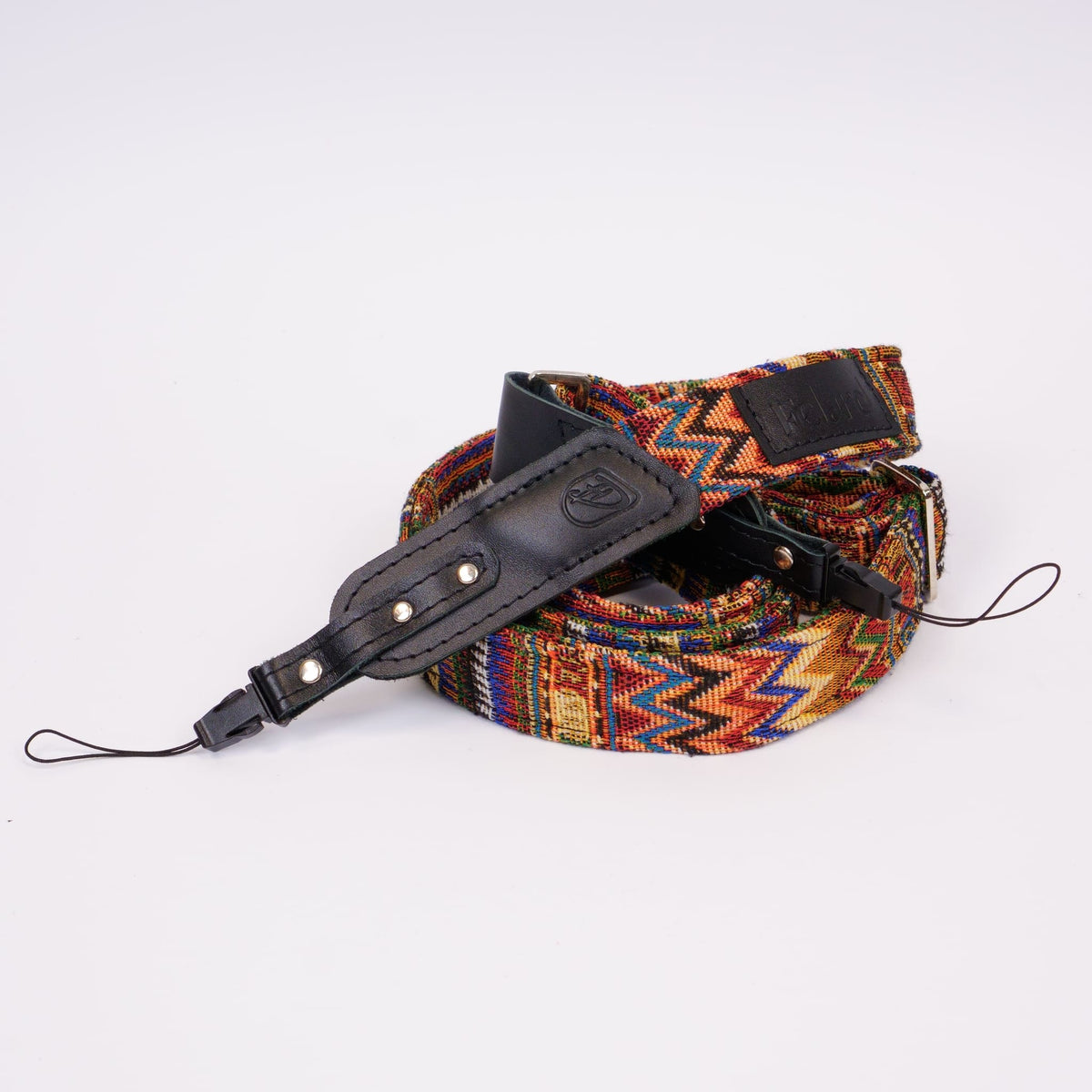 A coiled camera strap featuring a vibrant, multicolored geometric woven pattern with black leather accents and silver-tone metal rivets, photographed on a clean white background.