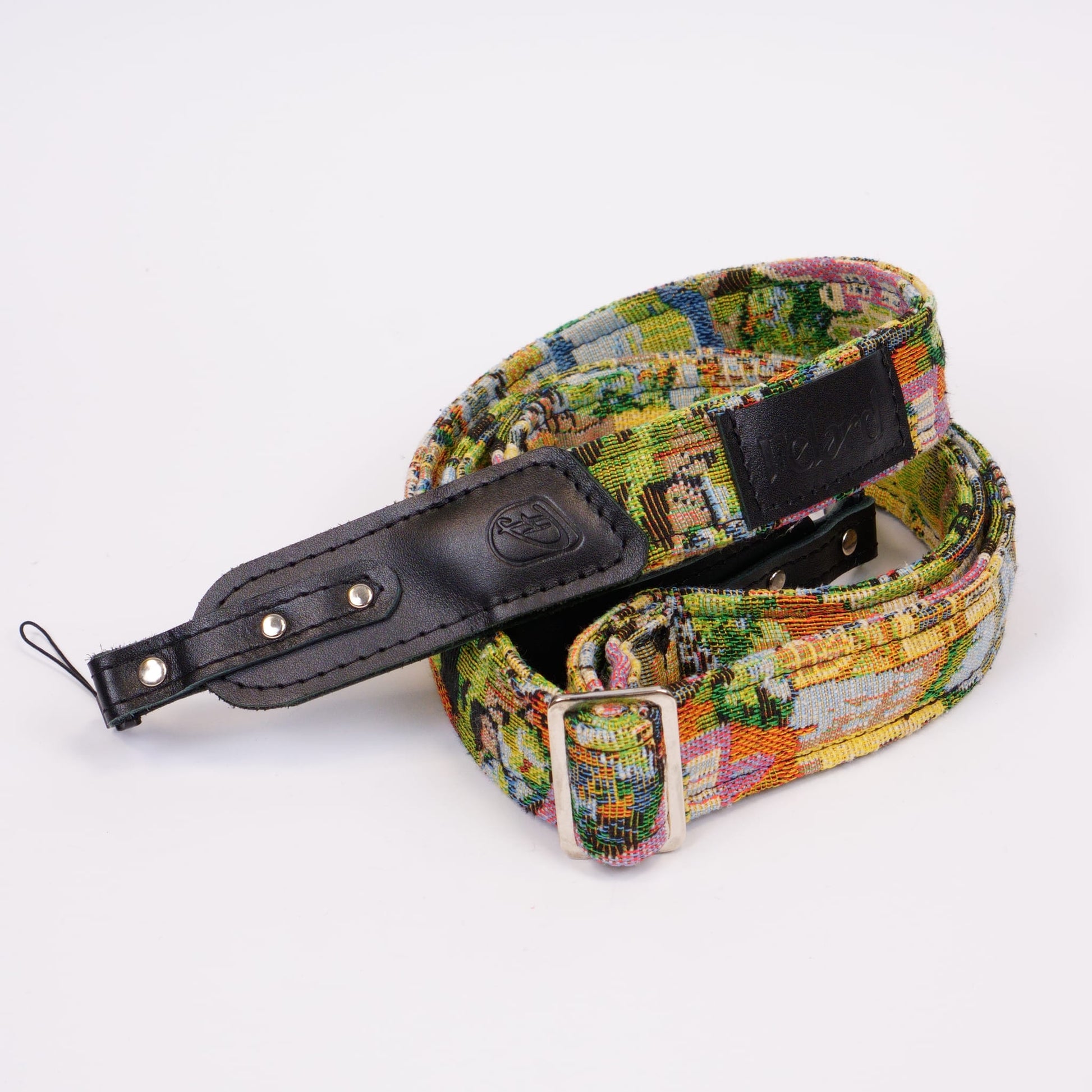 A woven canvas camera strap with a multicolor landscape pattern, featuring a black leather keeper and metal buckle, displayed on a clean white background.