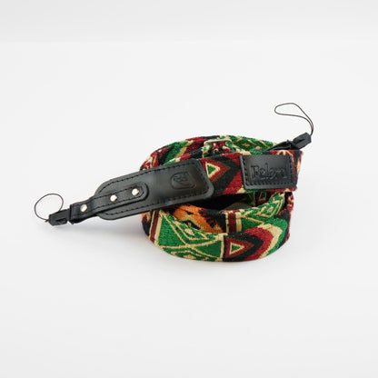 A woven camera strap with a vibrant green, red, and black geometric pattern, featuring black leather accents and silver-tone metal rings, displayed on a clean white background.
