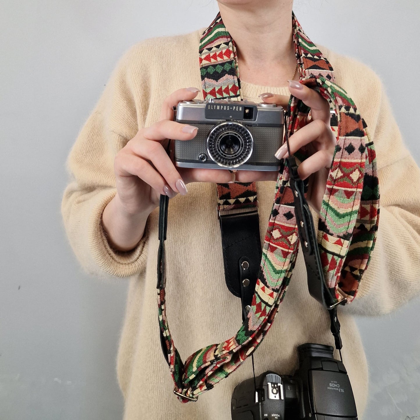 A person wearing a beige sweater holds a vintage silver camera with a colorful geometric patterned fabric camera strap. The strap features a Southwestern-style design with red, green, black, and beige patterns.