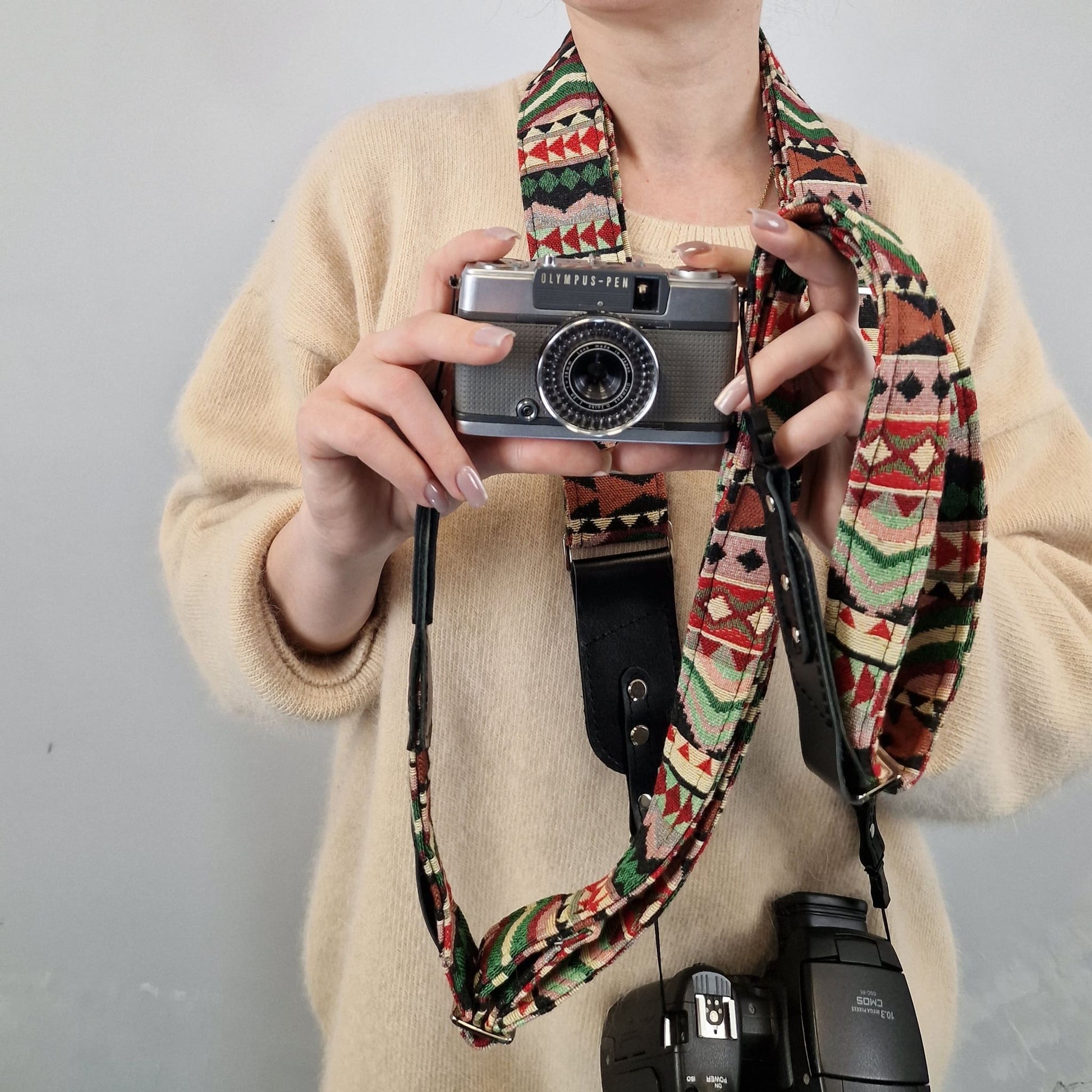 A person wearing a beige sweater holds a vintage silver camera with a colorful geometric patterned fabric camera strap. The strap features a Southwestern-style design with red, green, black, and beige patterns.
