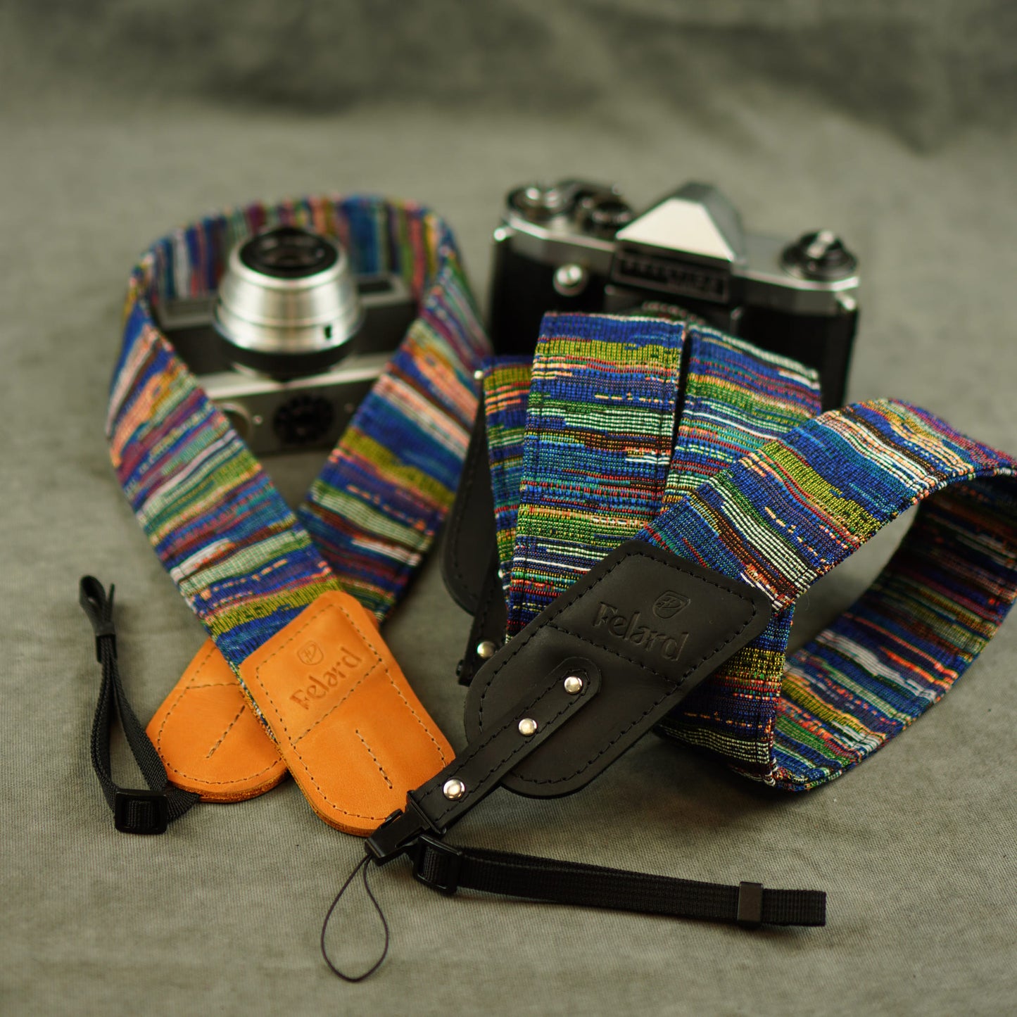 A multicolor woven camera strap with a black leather quick-release section and tan leather keeper, shown around a silver film camera on a gray textured background.