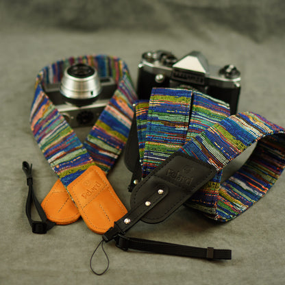A multicolor woven camera strap with a black leather quick-release section and tan leather keeper, shown around a silver film camera on a gray textured background.