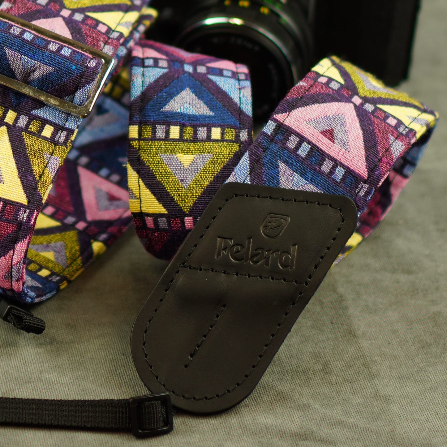A close-up of a colorful patterned camera strap with a black leather branded tag reading 'Felard'. The strap features a geometric design in pink, blue, yellow, and purple.