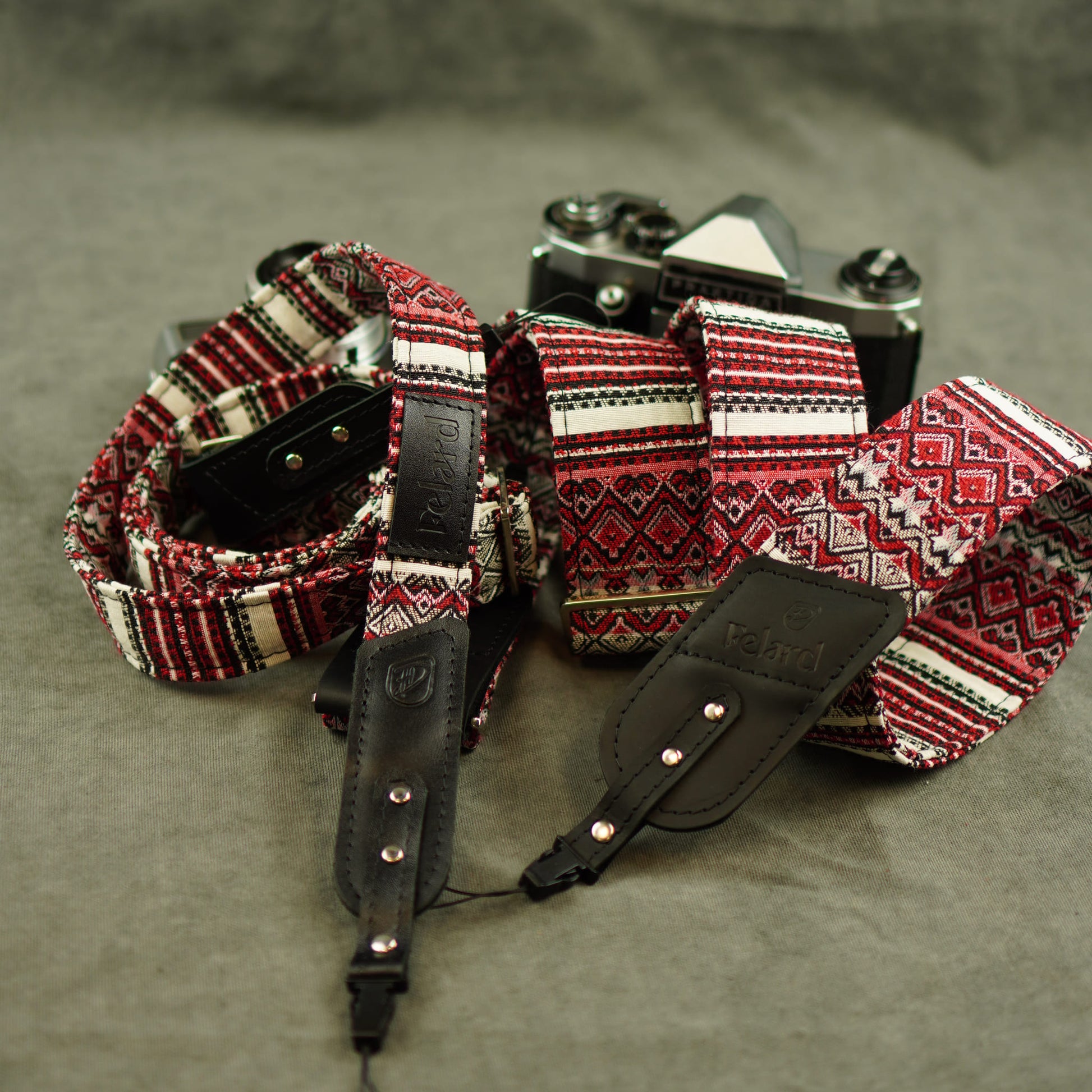A close-up of a camera strap with a red, black, and white geometric pattern and black leather accents, laid out on a gray textured surface with a vintage camera in the background.