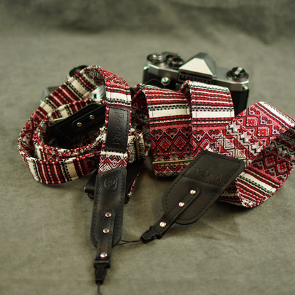 A close-up of a camera strap with a red, black, and white geometric pattern and black leather accents, laid out on a gray textured surface with a vintage camera in the background.