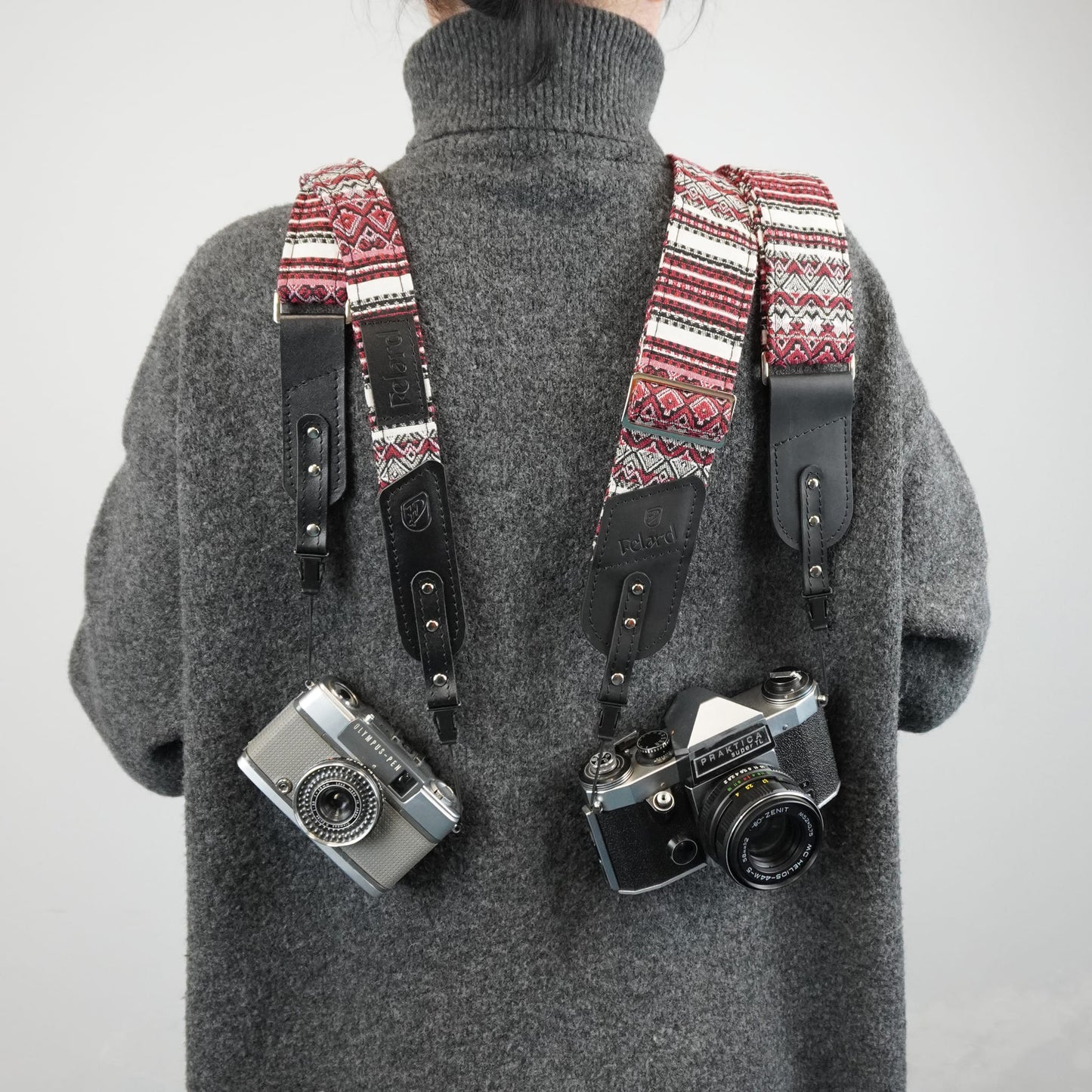 A person with a patterned camera strap system holding two cameras. The straps have a red, cream, and black geometric pattern with black leather sections.