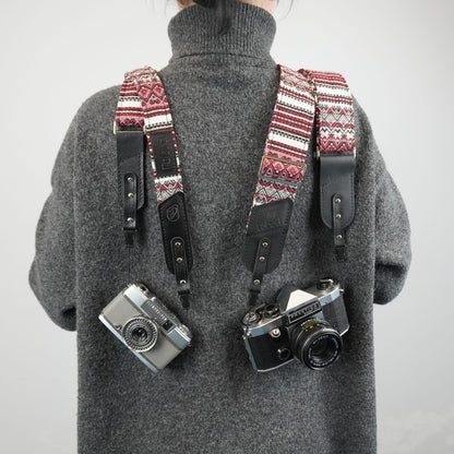 A person with a patterned camera strap system holding two cameras. The straps have a red, cream, and black geometric pattern with black leather sections.