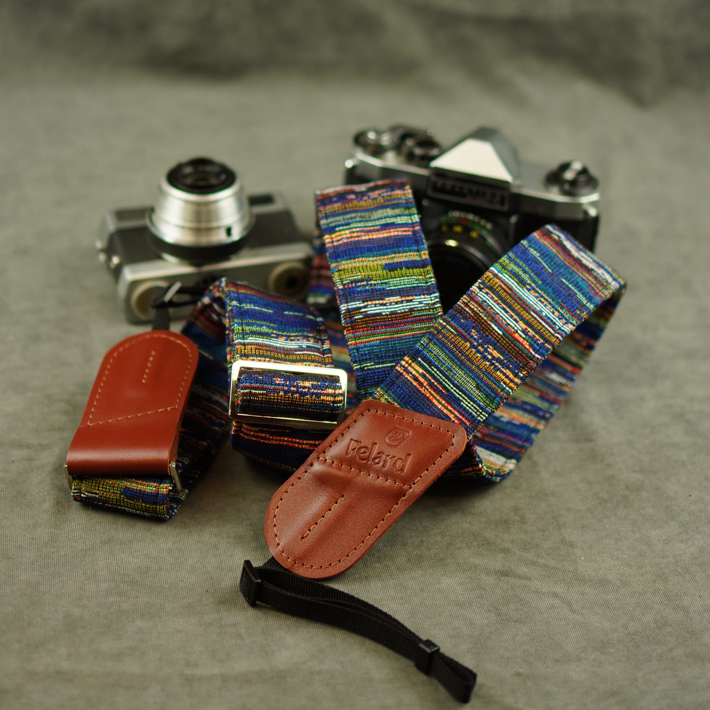 A multicolor woven camera strap with brown leather ends and a black wristlet loop, displayed with a vintage film camera and a brown leather pouch on a textured gray surface.