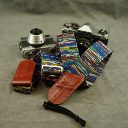 A multicolor woven camera strap with brown leather ends and a black wristlet loop, displayed with a vintage film camera and a brown leather pouch on a textured gray surface.