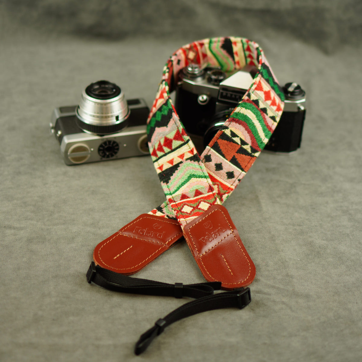 A patterned camera strap with red leather ends and a black adjustable section, displayed on a gray textured surface next to two vintage cameras.