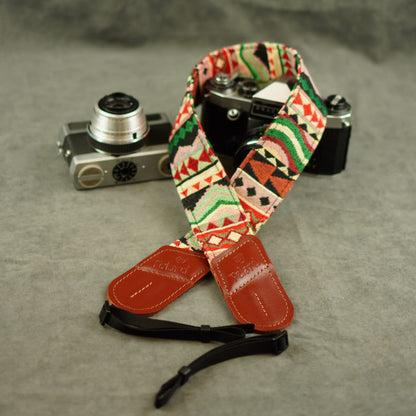 A patterned camera strap with red leather ends and a black adjustable section, displayed on a gray textured surface next to two vintage cameras.