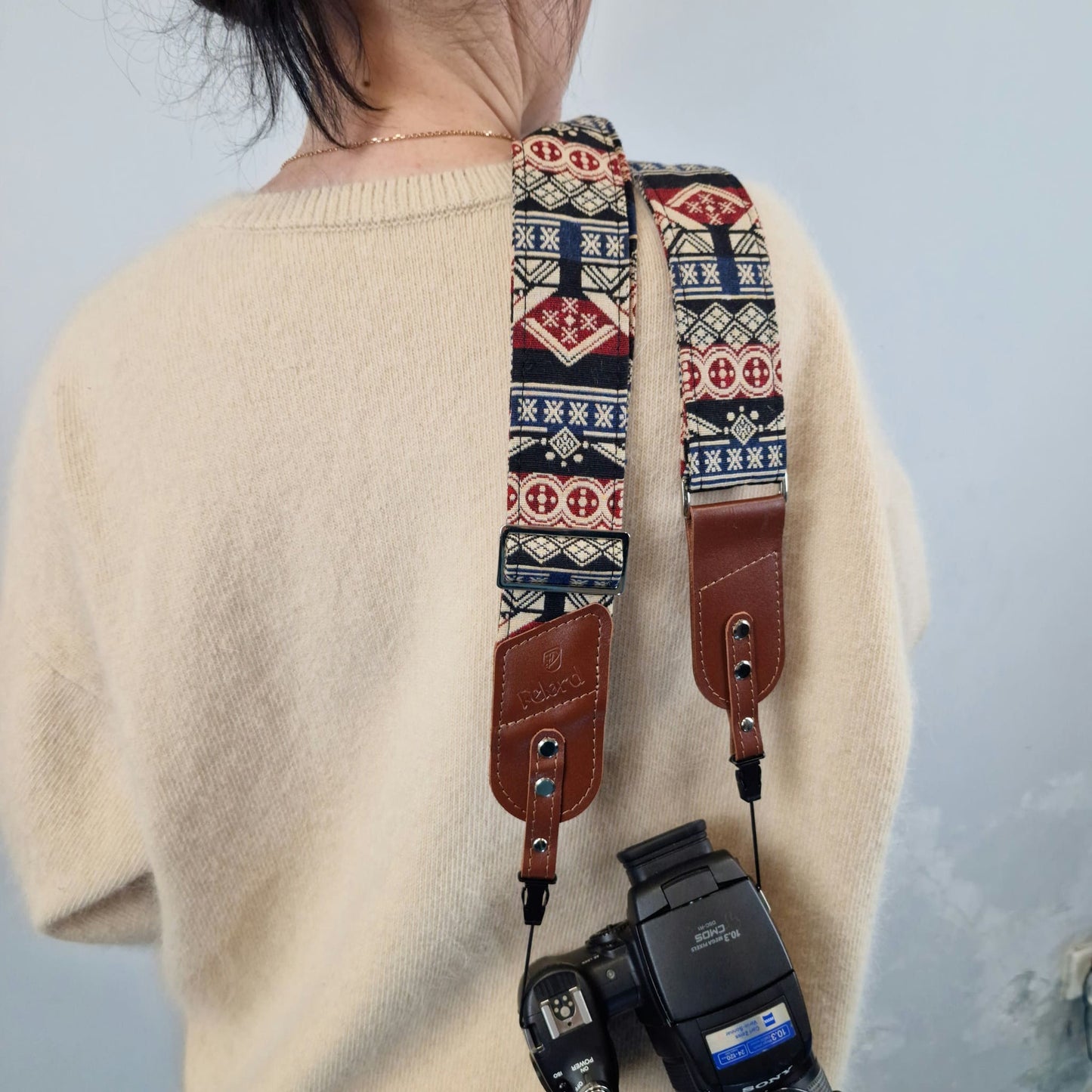 A person shown from behind, carrying a camera with a patterned camera strap. The strap features a geometric Southwestern-style design in navy blue, red, and beige, with brown leather accents.