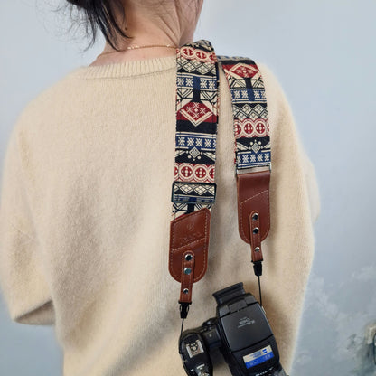 A person shown from behind, carrying a camera with a patterned camera strap. The strap features a geometric Southwestern-style design in navy blue, red, and beige, with brown leather accents.
