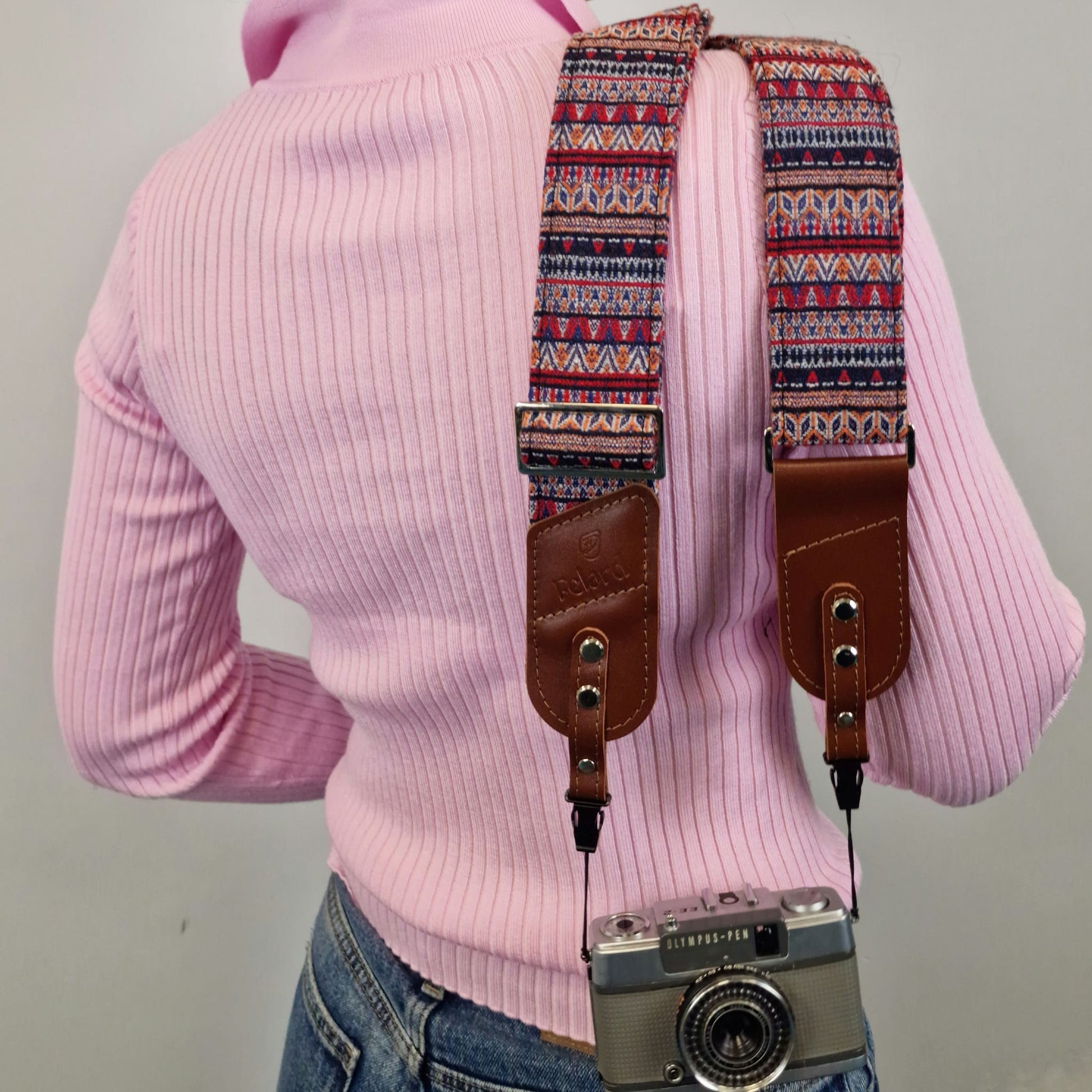 A person shown from the back while wearing a colorful geometric patterned camera strap with brown leather attachments. A vintage silver camera is hanging from the strap.