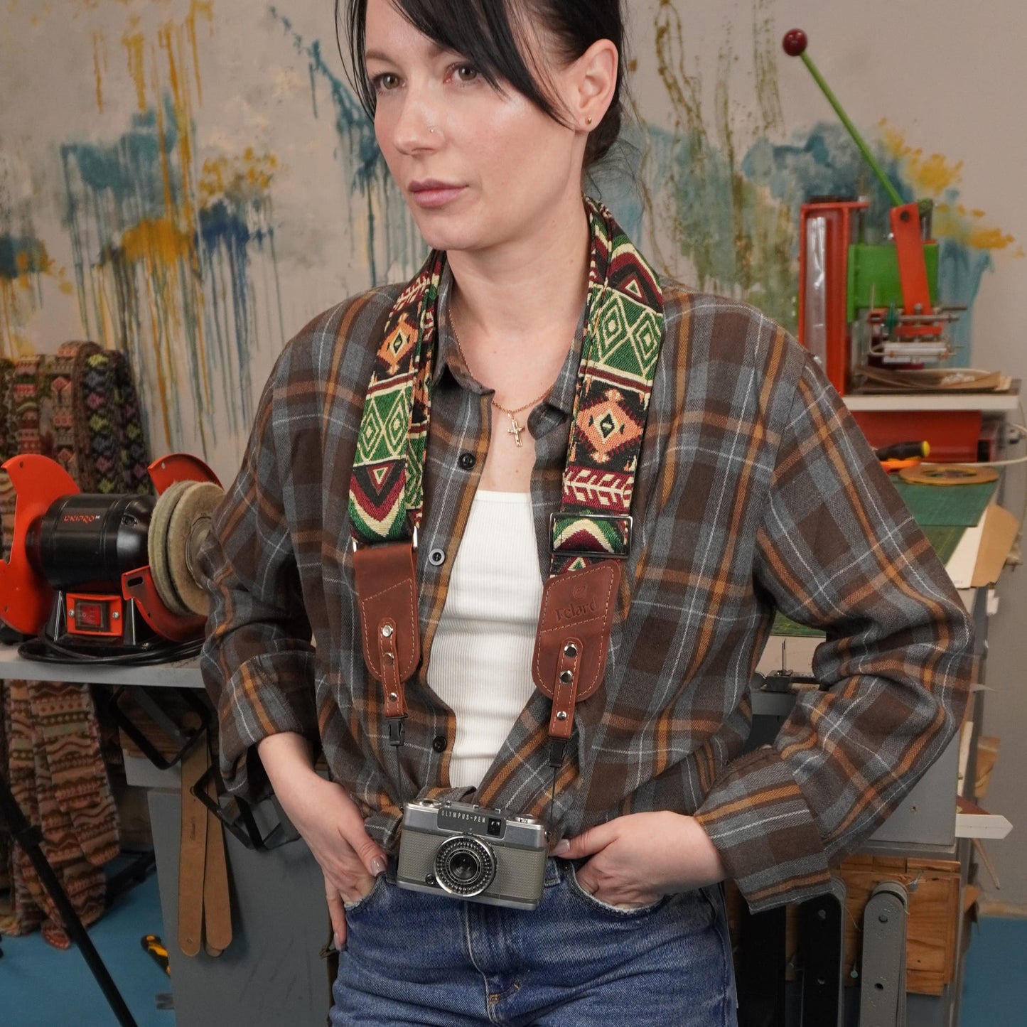 A woman with a colorful geometric patterned camera strap and brown leather, holding a vintage silver camera at her waist in a creative studio setting.