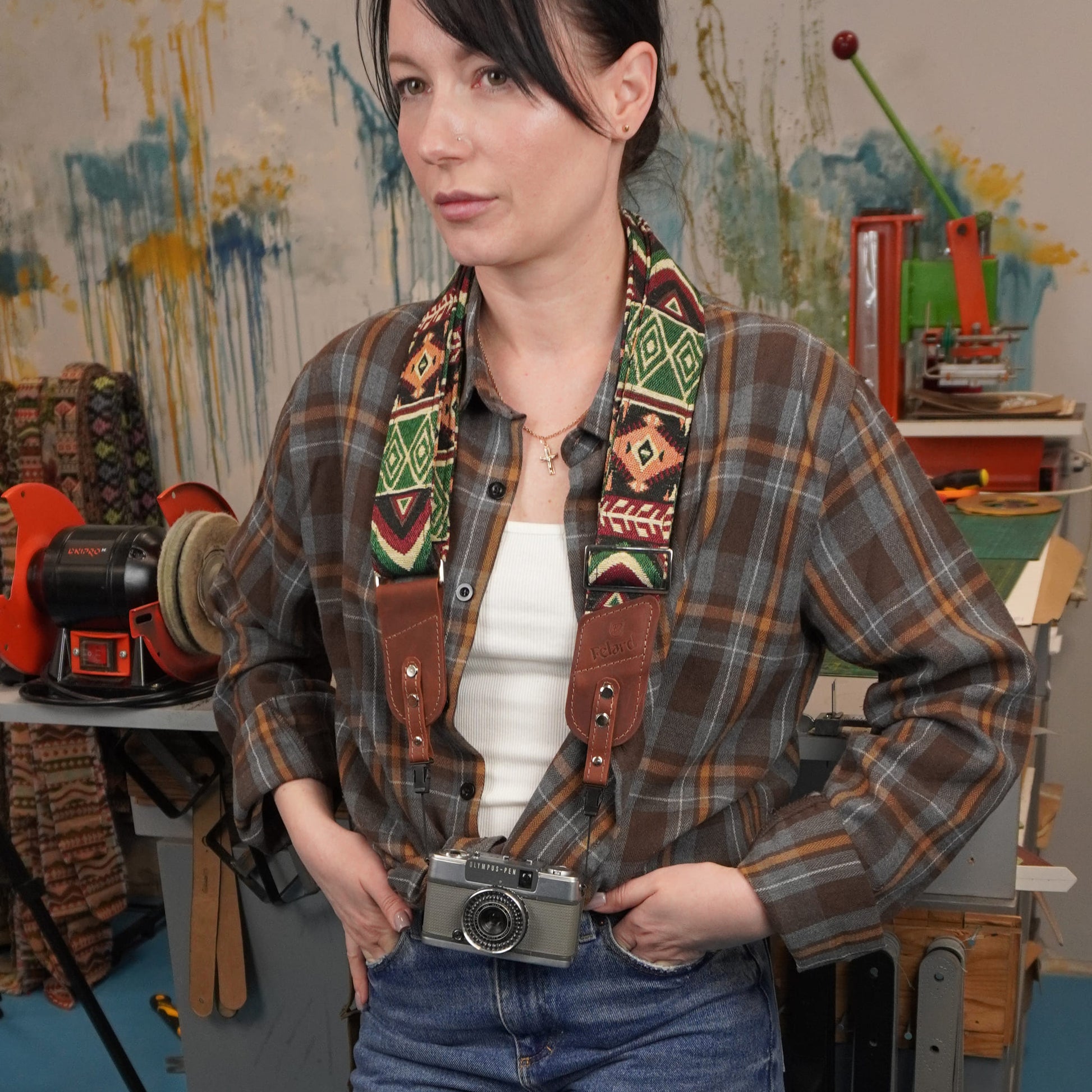 A woman with a colorful geometric patterned camera strap and brown leather, holding a vintage silver camera at her waist in a creative studio setting.