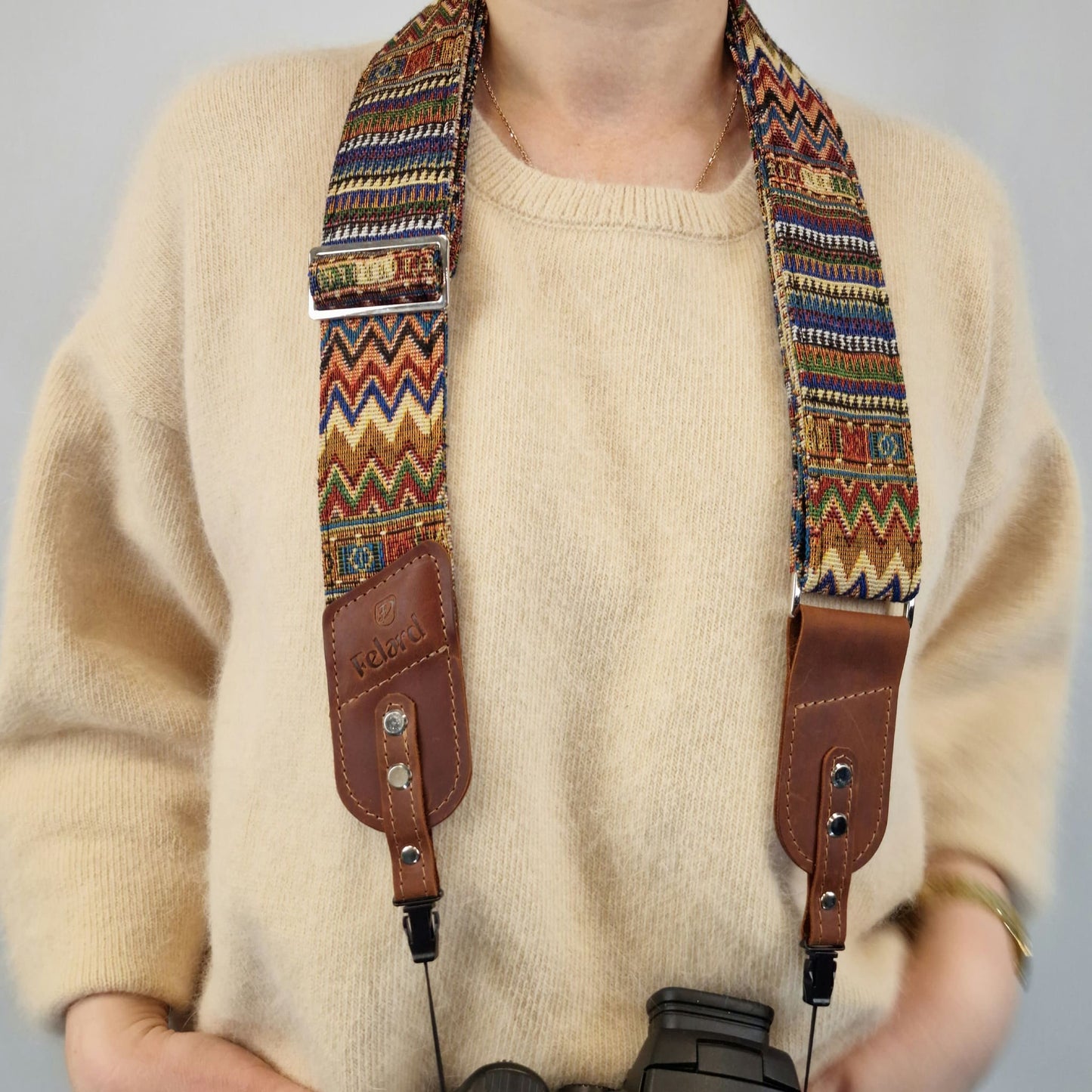A close-up of a person with patterned camera strap with brown leather accents. The strap has a multicolor geometric woven design and is shown holding a camera.