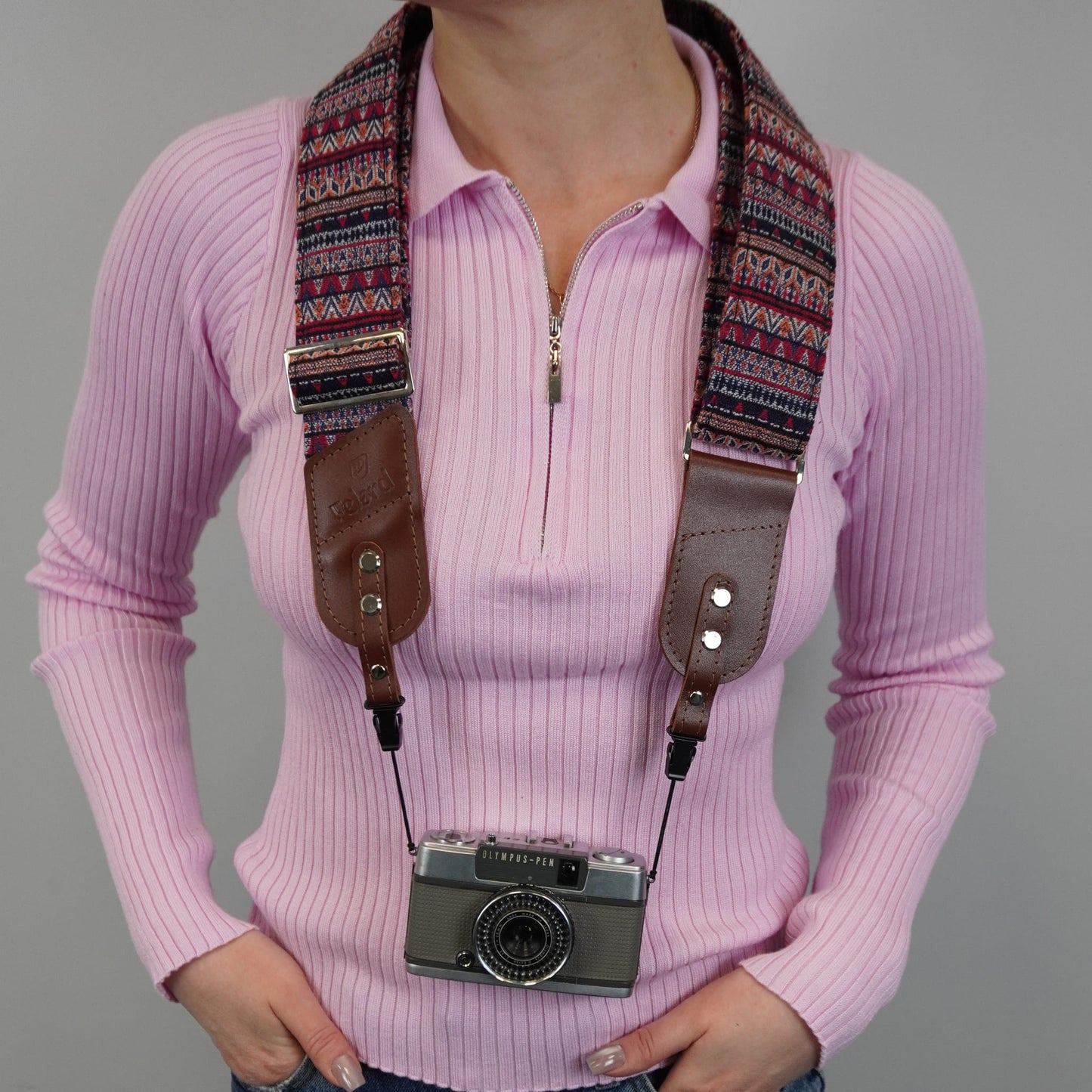 A person models a camera strap with brown leather side panels and a multicolor geometric patterned fabric. A vintage silver camera is hanging from the strap against a plain gray background.