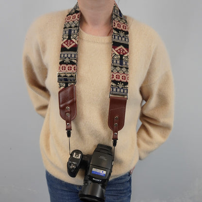 A person models a patterned camera strap with brown leather ends, holding a Sony camera with a flash. The camera strap features a geometric Southwestern-style pattern in red, beige, and black.
