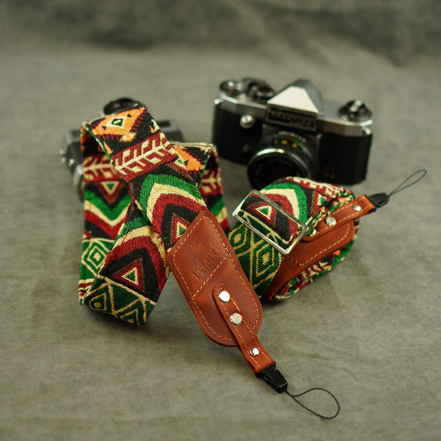 Camera strap with a colorful geometric pattern in red, green, black, and beige with brown leather. A vintage silver and black camera is placed behind it on a textured gray surface.