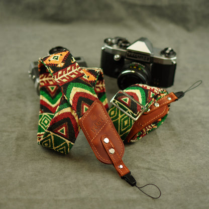 Camera strap with a colorful geometric pattern in red, green, black, and beige with brown leather. A vintage silver and black camera is placed behind it on a textured gray surface.