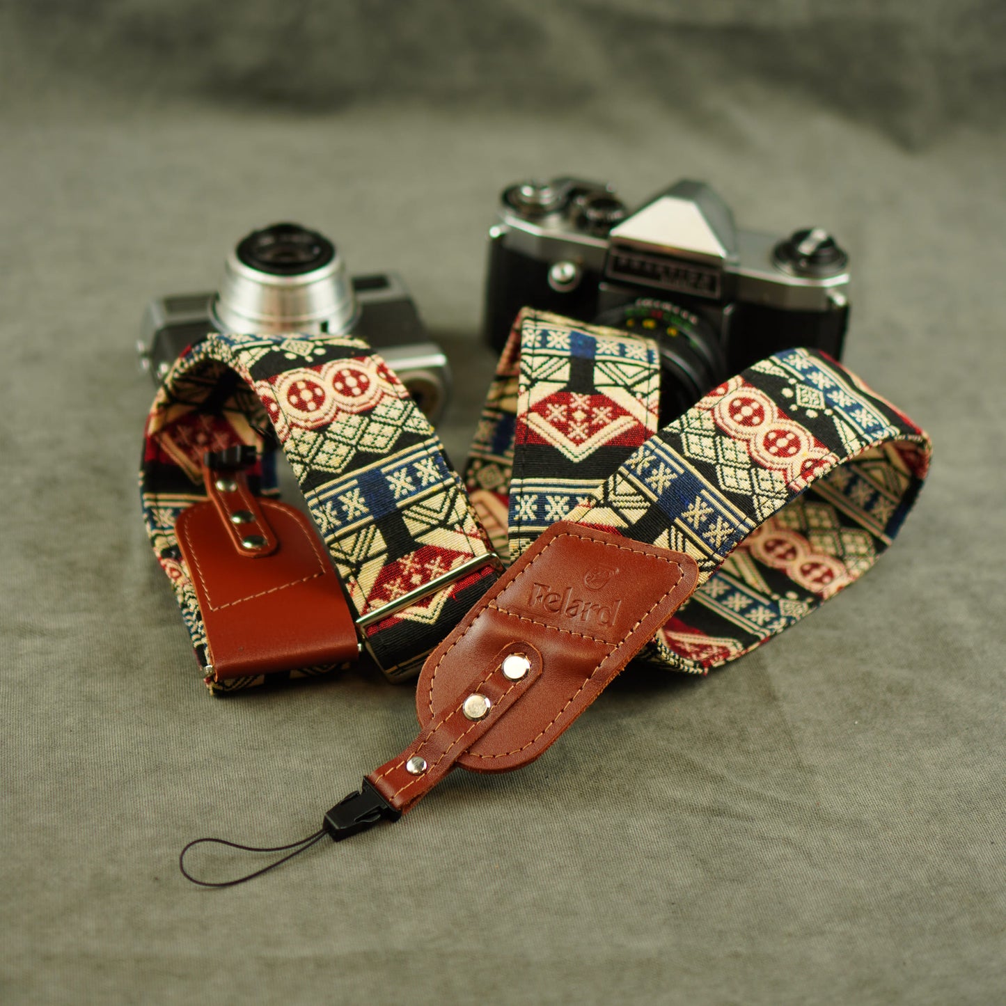 A patterned camera strap with brown leather accents and a small leather luggage tag, displayed in front of two vintage cameras on a gray textured background.
