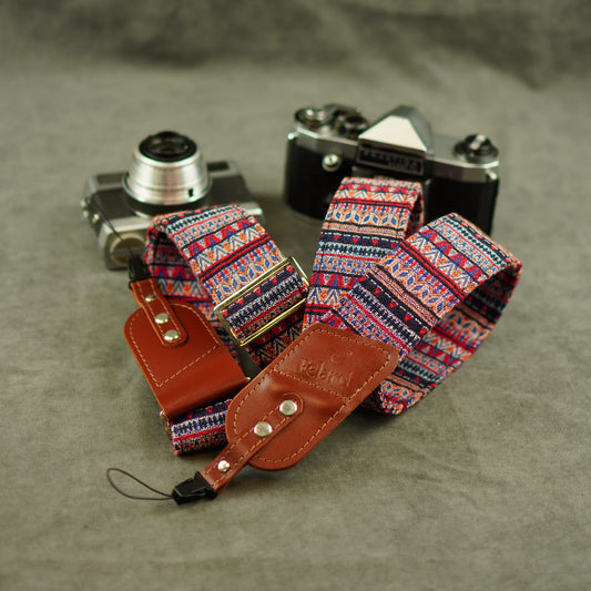 A camera strap set displayed on a textured gray surface, featuring a colorful geometric patterned fabric strap, a brown leather keeper, and two vintage cameras in the background.