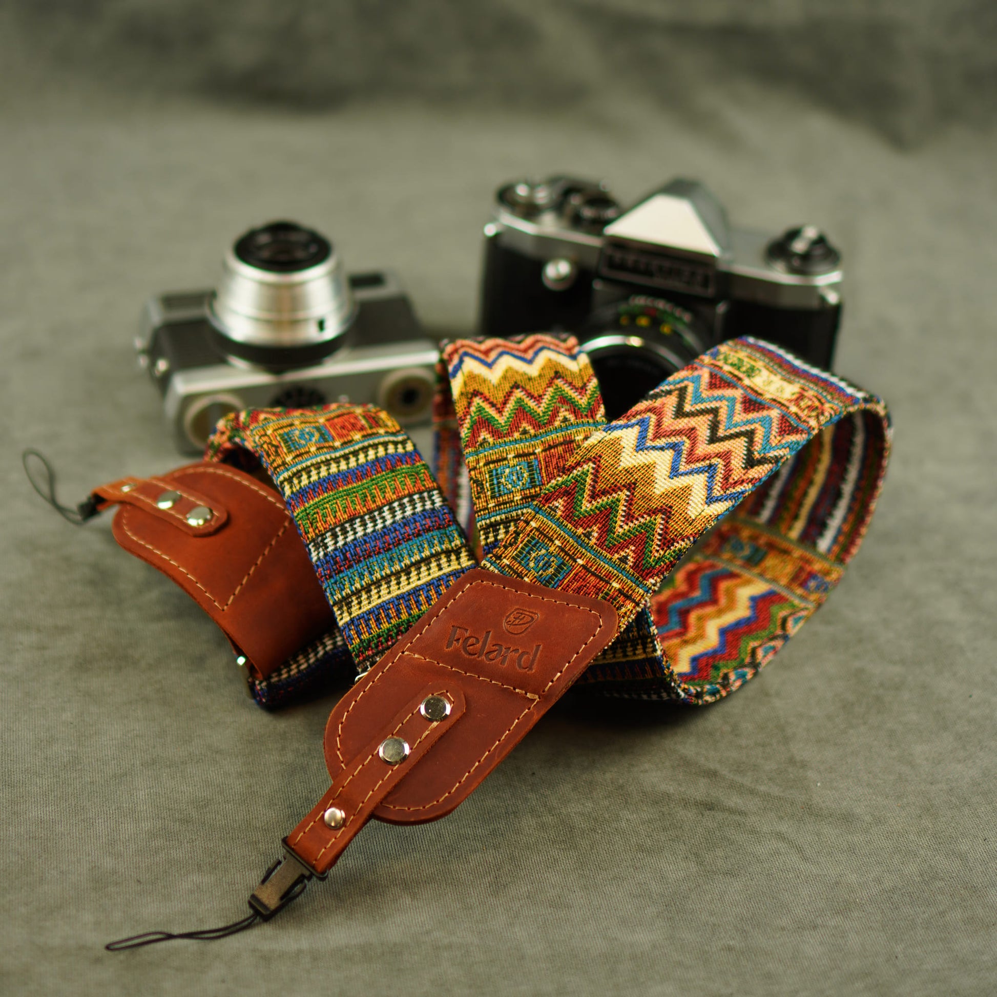 A multicolor patterned camera strap with brown leather ends and a visible Felard logo, laid over two vintage cameras on a gray textured surface.
