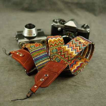 A multicolor patterned camera strap with brown leather ends and a visible Felard logo, laid over two vintage cameras on a gray textured surface.