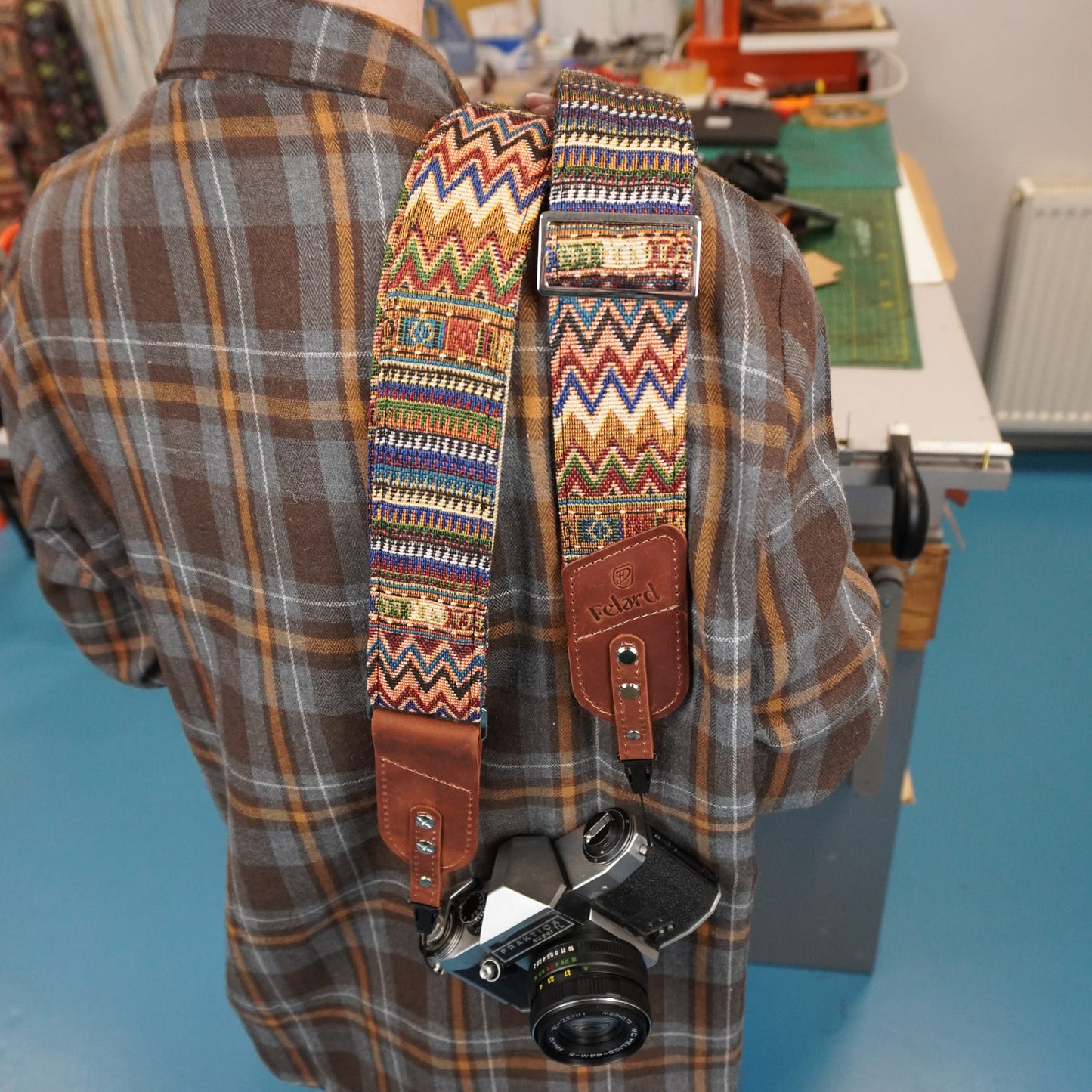 A person carrying a camera with a colorful patterned woven camera strap. The strap features brown leather ends and a central panel with geometric patterns in red, blue, yellow, and white.